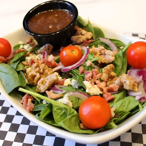 A spinach salad with cherry tomatoes, red onion, bacon, blue cheese, and crispy chicken, with dressing.