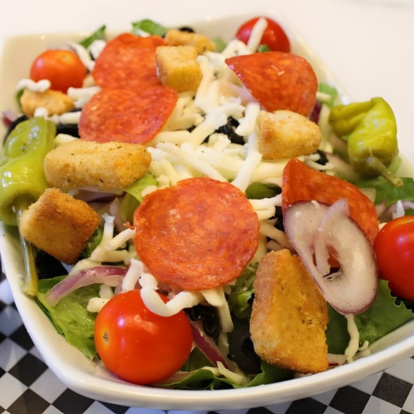 A pizza salad with pepperoni, cheese, tomatoes, croutons, and peppers in a white bowl.