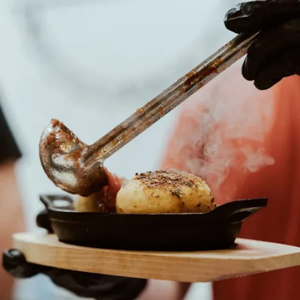 A gloved hand ladles sauce onto a steaming dish of Twisted Sisters Tap House arancini balls.