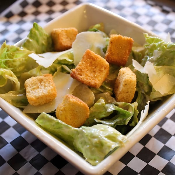 Caesar salad with croutons and parmesan cheese in a white bowl on a checkered tablecloth.