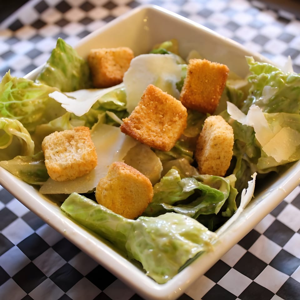 Caesar salad with croutons and parmesan cheese in a white bowl on a checkered tablecloth.