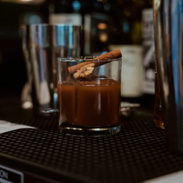A short glass of amber liquid, garnished with a cinnamon stick and a walnut, on a dark bar.