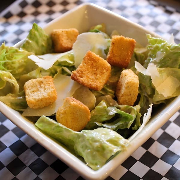 Caesar salad with crisp lettuce, croutons, and parmesan cheese in a white bowl on a checkered tablecloth.